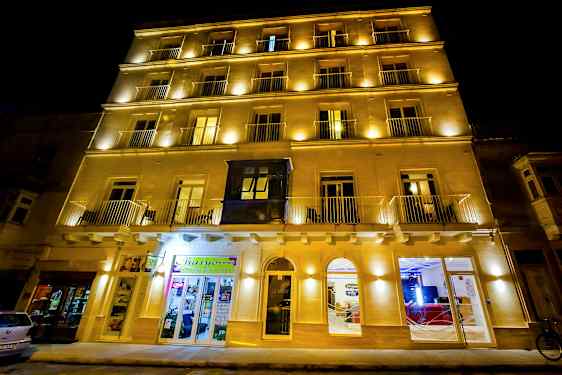 En bygning fotografert om natten med opplyste fasader og flere vinduer. Noen butikker og innganger er synlige. (AI-generert tekst)