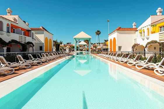 Et utendørsbasseng omgitt av solsenger og fargerike bygninger med en klar himmel. (AI-generert tekst)