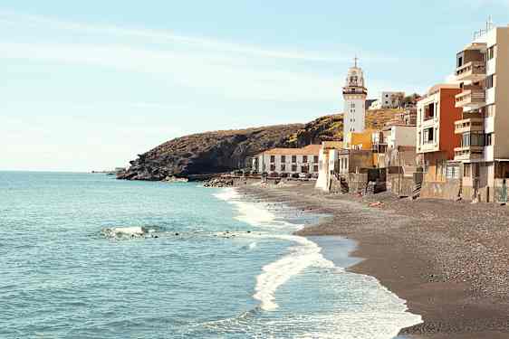 Bilde av en kyst med svarte sandstrender, et fyrtårn og bygninger ved vannkanten. (AI-generert tekst)