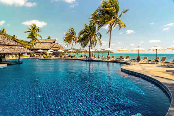 Bilde av et utendørsbasseng omgitt av solsenger og palmer, med havet i bakgrunnen. (AI-generert tekst)