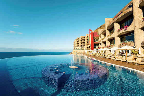 Et infinitybasseng ved strandkanten med solsenger og hotellbygninger i bakgrunnen. (AI-generert tekst)