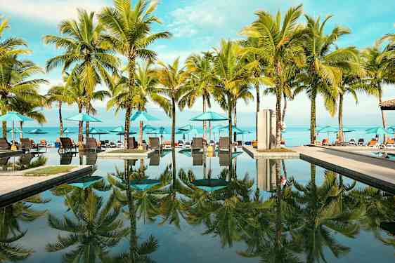 Utendørs basseng med palmer og solsenger ved stranden. (AI-generert tekst)