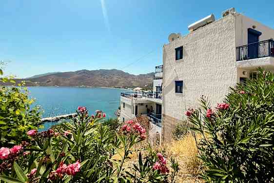 Hvit bygning med blå detaljer ved havet, omgitt av busker med rosa blomster. (AI-generert tekst)