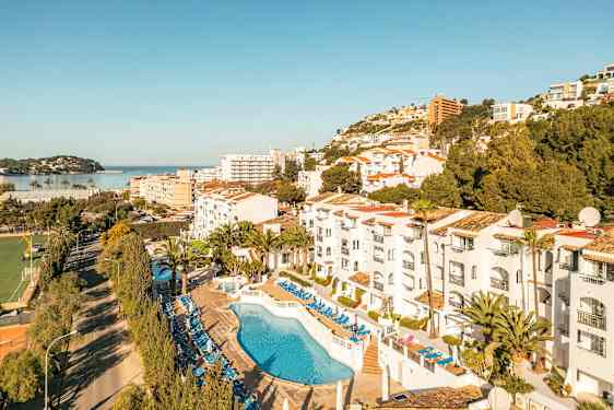 Bilde av hotellområde med basseng og solsenger, omgivelser med trær og bygninger samt en utsikt mot havet. (AI-generert tekst)