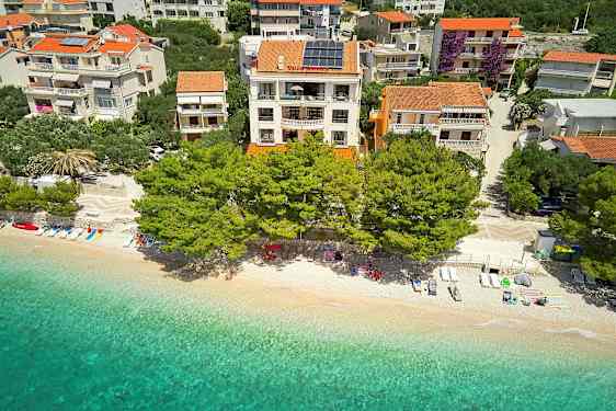 Flyfoto av en strand med solsenger, parasoller og klart vann foran bygninger med terrakottatak omgitt av trær. (AI-generert tekst)