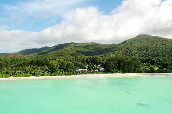 En strand med hvit sand foran en skogkledd fjellkjede og turkis vann. (AI-generert tekst)