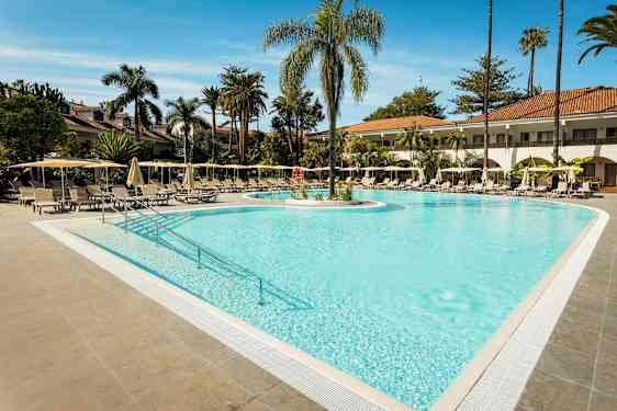 Et utendørs basseng omgitt av solsenger og parasoller med palmetrær i bakgrunnen. (AI-generert tekst)