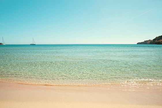 Strand med klart vann og to seilbåter i havet. (AI-generert tekst)