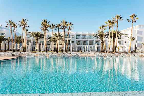 Hotellets bassengområde med klart blått vann, omgitt av solsenger, parasoller og palmer. I bakgrunnen sees en hvit bygning med balkonger. (AI-generert tekst)