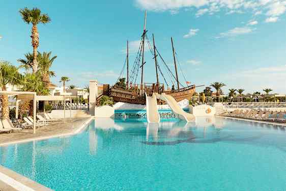 Utendørsbasseng med sklier fra et sjørøverskip og omkringliggende solsenger. (AI-generert tekst)