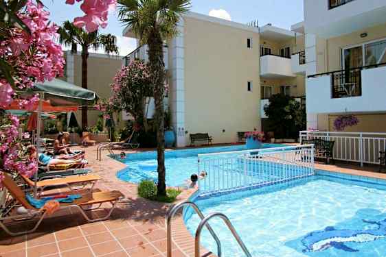 Et svømmebasseng med solsenger, parasoller og blomster rundt, med bygninger i bakgrunnen. (AI-generert tekst)