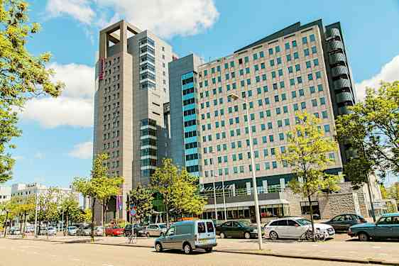 En moderne bygning med flere etasjer omgitt av parkerte biler og trær. (AI-generert tekst)