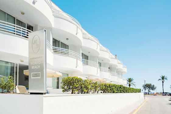 Den hvite bygningen tilhører Morito Beach Apartments, med palmer og sjø i bakgrunnen. (AI-generert tekst)