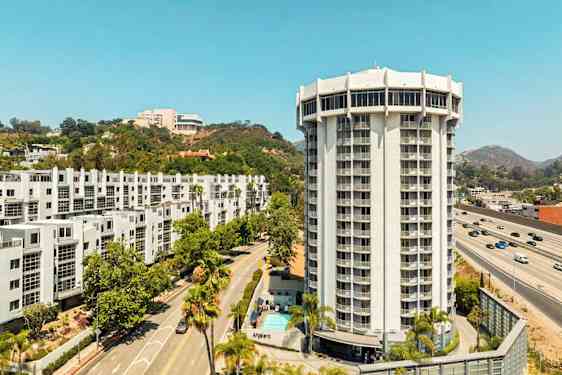 En moderne høyblokk omgitt av veier, grøntområder og andre bygninger. (AI-generert tekst)