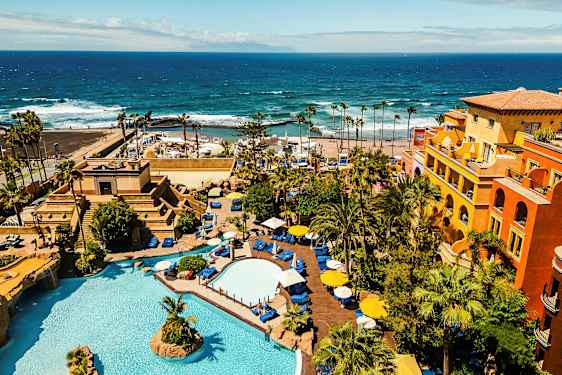 Et hotellområde med bassenger, vegetasjon og fargerike bygninger ved havet. (AI-generert tekst)