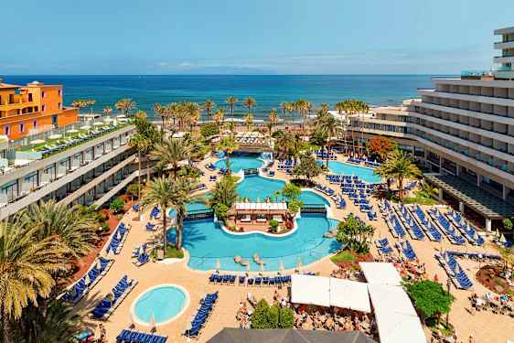 Det er en utsikt over et hotellområde med flere bassenger, omgitt av solsenger og parasoller, samt trær og bygninger. I bakgrunnen er havet synlig. (AI-generert tekst)