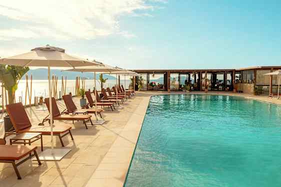 Bildet viser et utendørs svømmebasseng med solsenger og parasoller langs bassengkanten, med havet og en skyet himmel i bakgrunnen. (AI-generert tekst)