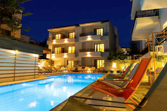 Bildet viser en opplyst hotellbygning og et utendørs basseng omgitt av solsenger og lounge-stoler på kveldstid. (AI-generert tekst)