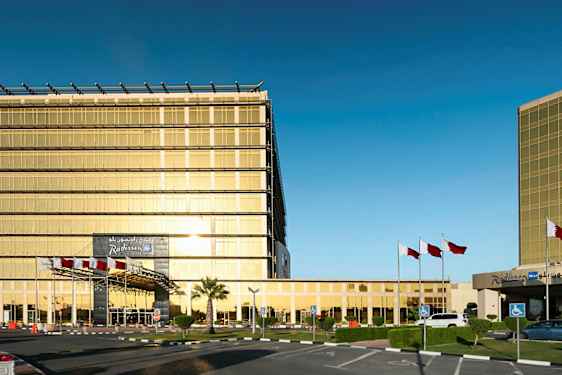 En moderne bygning med gullfargede glassvegger, omgitt av flaggstenger og en parkeringsplass. (AI-generert tekst)