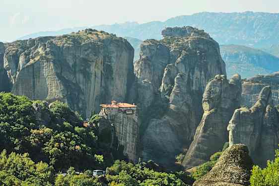 Et kloster plassert på en klippeformasjon omgitt av grønne skoger og fjell. (AI-generert tekst)
