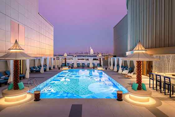 En takterrasse med et dekorativt basseng omgitt av solsenger og små parasoller. I bakgrunnen sees Burj Al Arab og Dubais skyline under en lilla kveldshimmel. (AI-generert tekst)