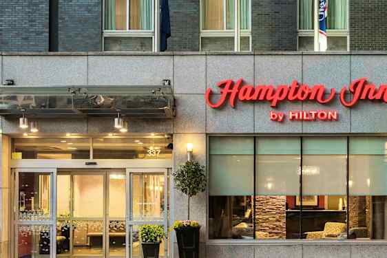 Hotellinngang med skilt "Hampton Inn by Hilton" over dørene. (AI-generert tekst)