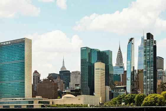 Bildet viser en bysilhuett med flere høye bygninger og skyskrapere omgitt av grøntområder under en klar himmel. (AI-generert tekst)