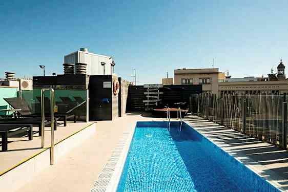 Et utendørs basseng på en takterrasse omgitt av solsenger og bygninger i bakgrunnen. (AI-generert tekst)