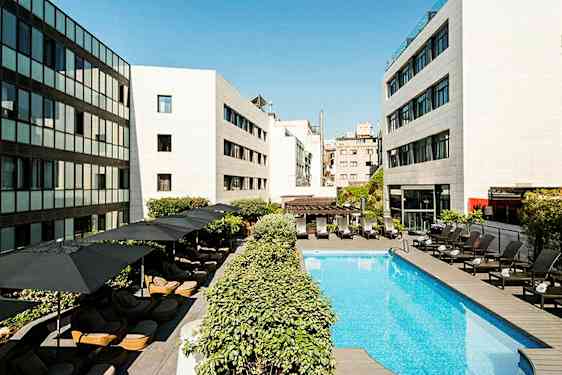 Et basseng på et hotell omgitt av solsenger og parasoller, med moderne bygninger rundt. (AI-generert tekst)