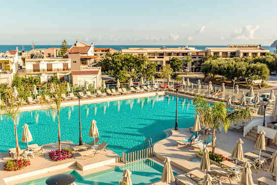 Et utendørs svømmebasseng omgitt av solsenger og parasoller, med bygninger og havet i bakgrunnen. (AI-generert tekst)