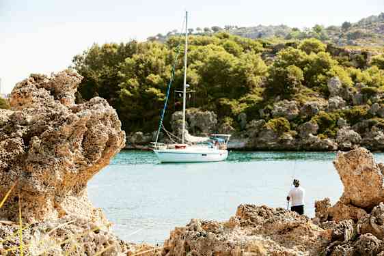 En seilbåt ligger fortøyd ved en fredelig vik omgitt av klipper og skog, med en person som fisker fra stranden. (AI-generert tekst)