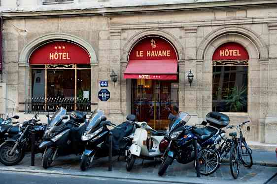 Bilde som viser inngangen til et hotell med navnet Le Havane, tre stjerner vises på markisen. Foran bygningen står mopeder og sykler parkert. (AI-generert tekst)