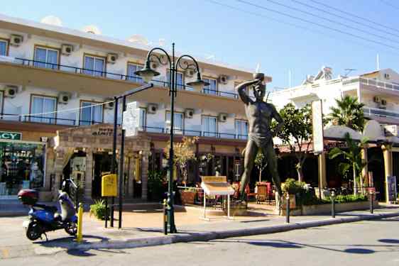 Statue foran en bygning med butikker og restaurant. En scooter er parkert ved siden av. (AI-generert tekst)