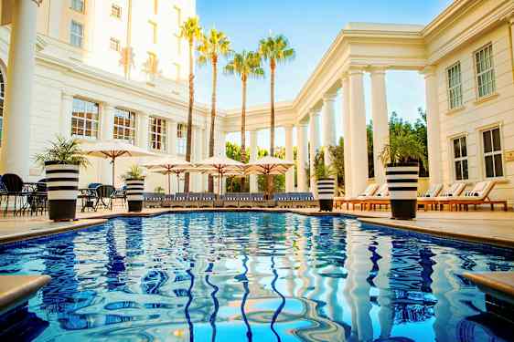 Bassengområde med skinnende blått vann, solsenger og parasoller omgitt av søyler og palmer. (AI-generert tekst)