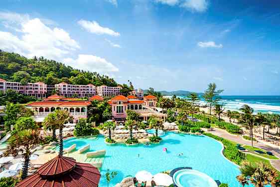 Bilde av et luksuriøst hotellområde ved havet, med et stort basseng omgitt av palmer og bygninger med røde tak. (AI-generert tekst)