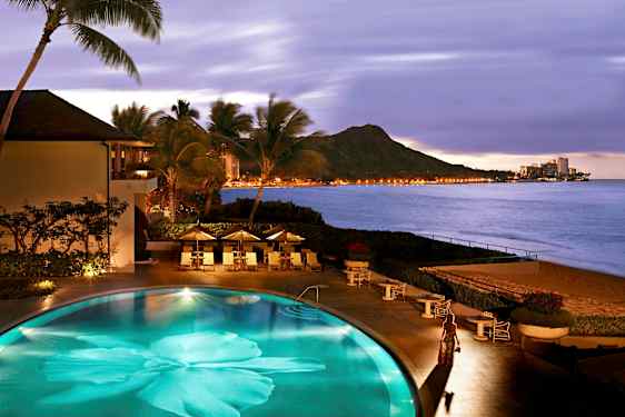 Et basseng ved stranden omgitt av palmer, med utsikt over fjell og lysene fra en by ved sjøen. (AI-generert tekst)