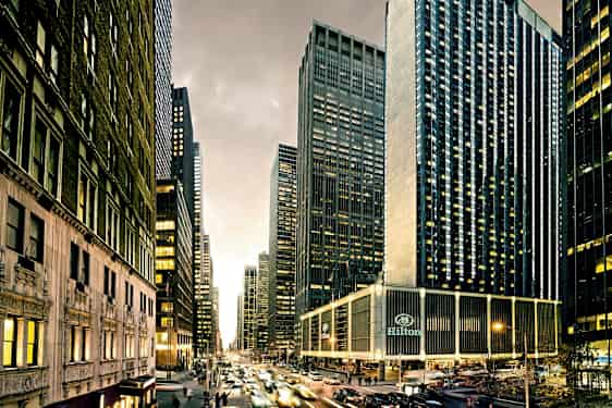 En byutsikt med høye skyskrapere, livlig trafikk og bygninger med mange vinduer. Hilton-hotellet vises nederst. (AI-generert tekst)