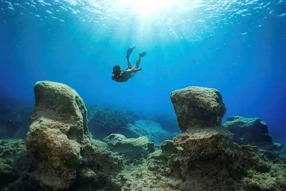 En snorkler svømmer under vann mellom store steiner med sollys som filtrerer ned fra overflaten. (AI-generert tekst)