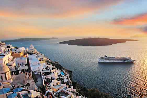 Hvite kalkede bygninger på en klippe over havet med et cruiseskip i bakgrunnen under en fargerik solnedgang. (AI-generert tekst)
