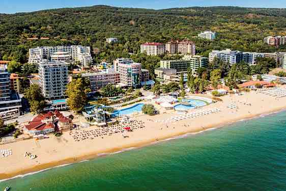 Flyfoto av en strand med solsenger og parasoller, et bassenganlegg og hoteller omgitt av grønne områder. (AI-generert tekst)