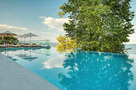 Et infinity-basseng med solsenger og parasoller ved siden av, omgitt av trær og med utsikt over havet. (AI-generert tekst)