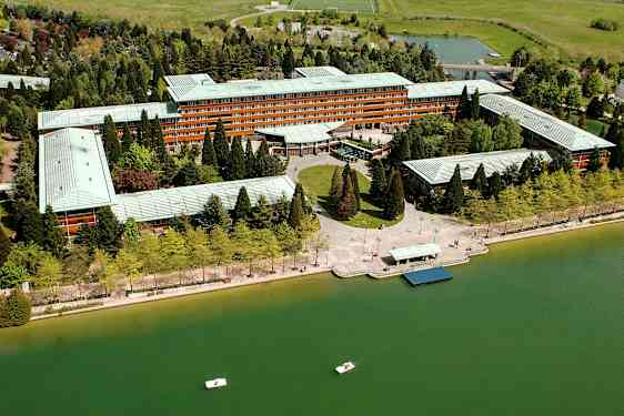 Flyfoto av en stor bygning omgitt av grønne trær ved en vannflate med små båter. (AI-generert tekst)