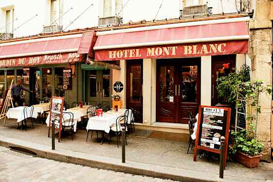 Utenfor et hotellbygg med restaurant hvor bord og stoler er satt opp på fortauet. (AI-generert tekst)