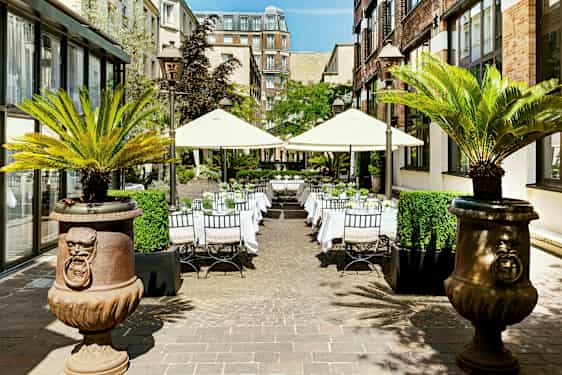 Utendørs selskapsområde med bord og parasoller omgitt av bygninger og palmer. (AI-generert tekst)