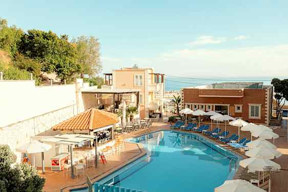 Et hotellbasseng med solsenger og parasoller ved siden av, samt bygninger og grøntområder i bakgrunnen. (AI-generert tekst)