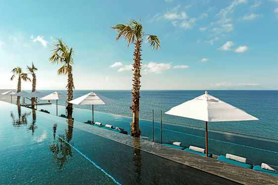 Et infinity-basseng med utsikt mot havet, omgitt av parasoller og palmer under en klar blå himmel. (AI-generert tekst)