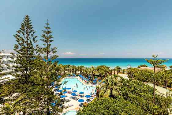 Bilde av hotellområde med basseng og solsenger omgitt av grøntområder og utsikt mot havet. (AI-generert tekst)