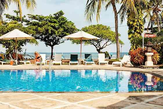 Bilde av et basseng med klart blått vann, omgitt av solsenger og parasoller med havet og palmetrær i bakgrunnen. (AI-generert tekst)