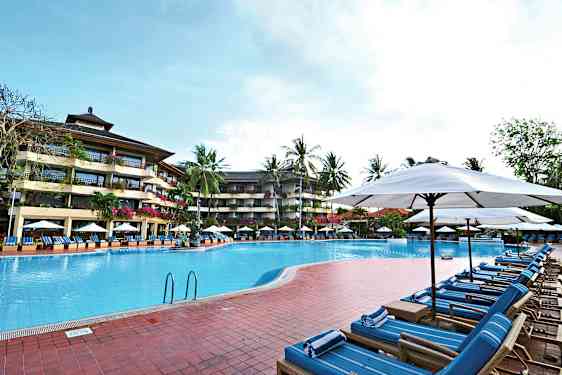 Bilde av et utendørs svømmebasseng omgitt av solsenger og parasoller, med et fleretasjes hotell og palmer i bakgrunnen. (AI-generert tekst)