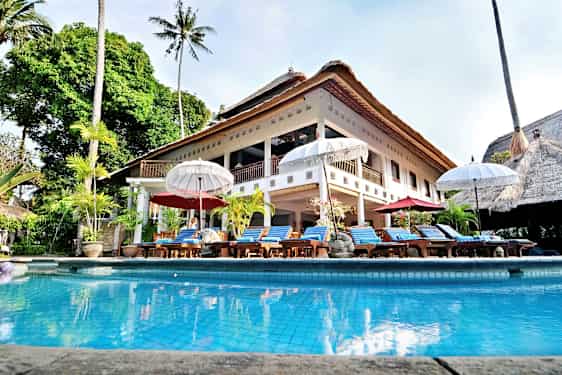 En stor villa med stråtak og et basseng foran, omgitt av solsenger og parasoller. (AI-generert tekst)